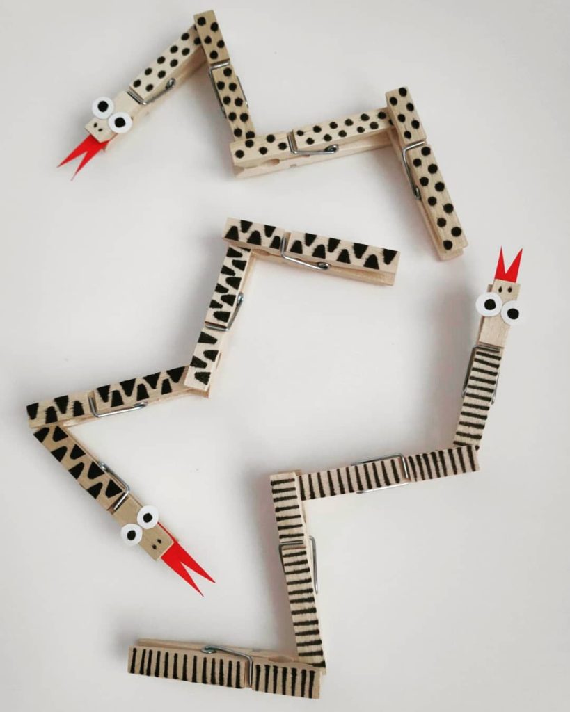 Flexible snakes crafted from wooden clothespins painted with patterns like stripes and dots, featuring googly eyes and red paper tongues, arranged in zigzag poses.

