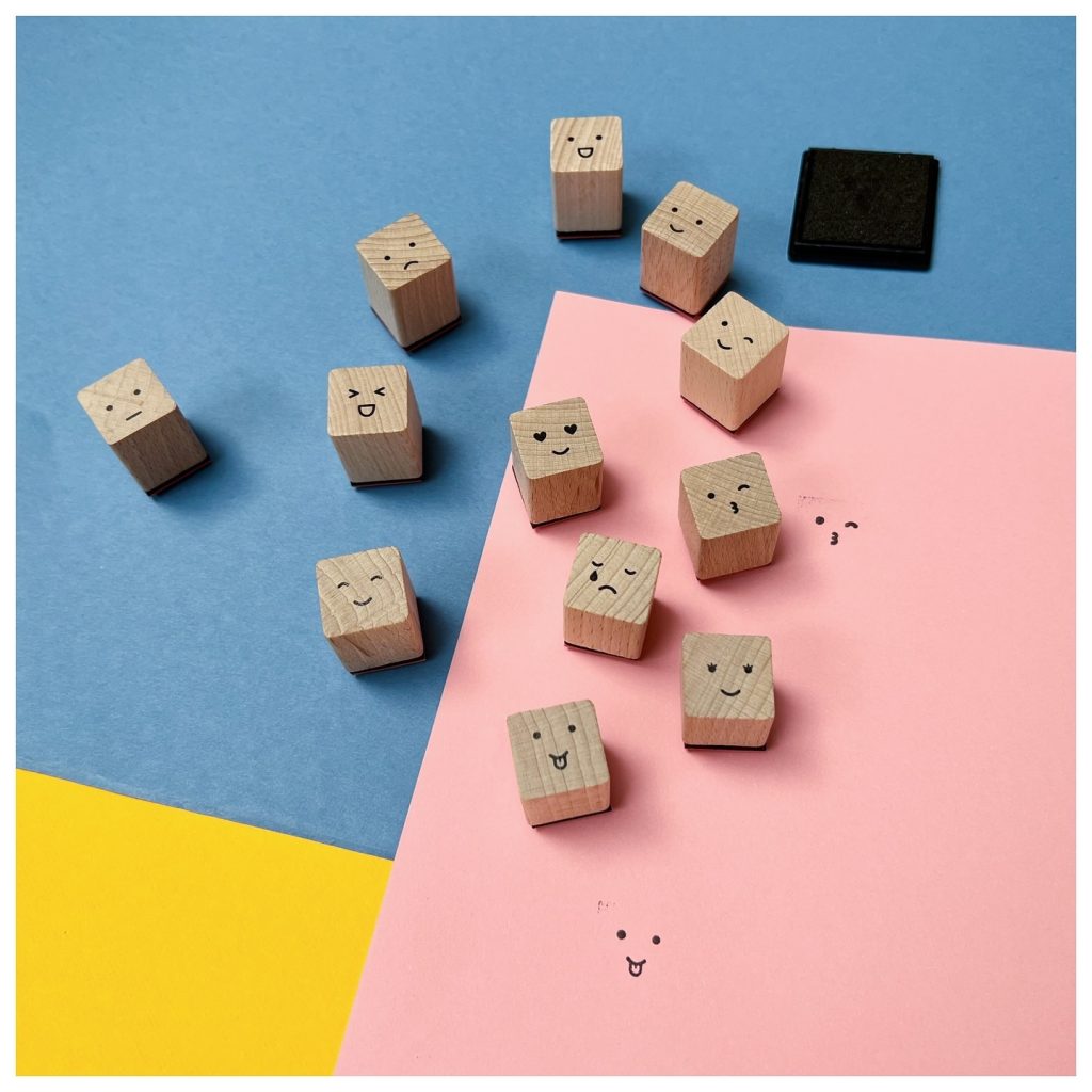 Wooden blocks decorated with various emoji-style faces, paired with a black ink pad, scattered on colorful paper surfaces in blue, pink, and yellow.