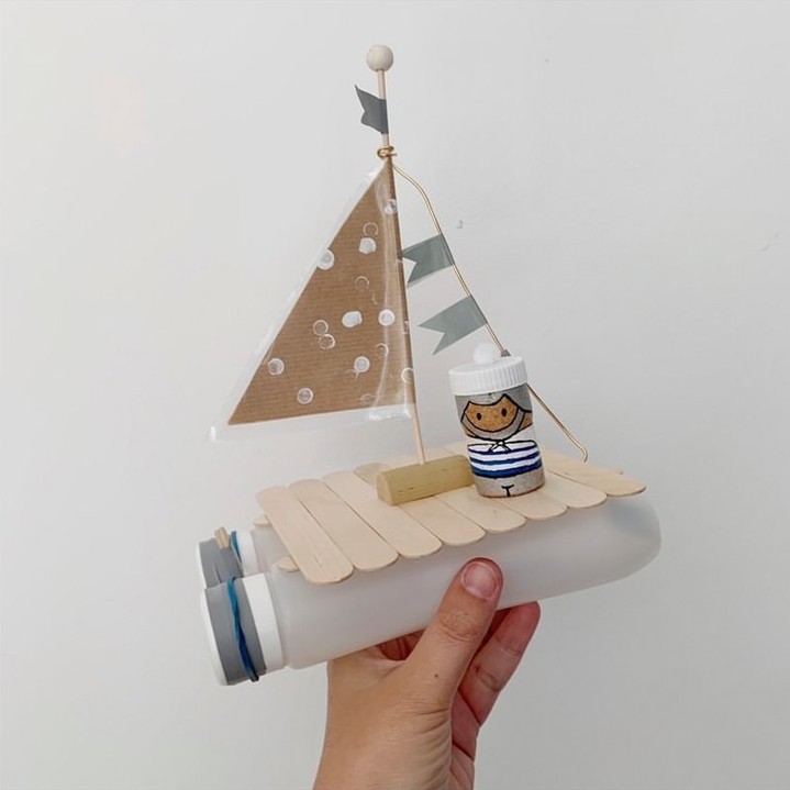 A sailboat crafted from a recycled plastic bottle with a wooden stick deck, a cardboard sail, and a cork sailor, decorated with small flags, held in a hand.






