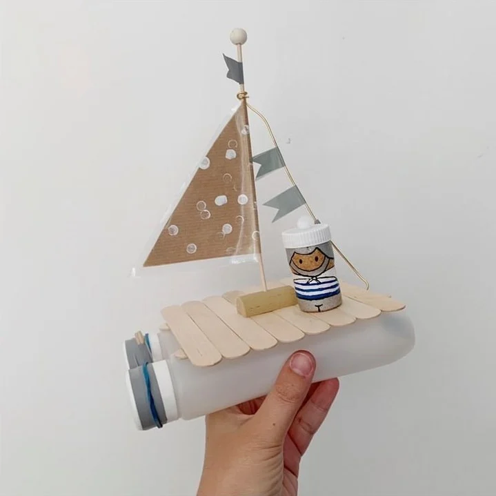 A sailboat crafted from a recycled plastic bottle with a wooden stick deck, a cardboard sail, and a cork sailor, decorated with small flags, held in a hand.