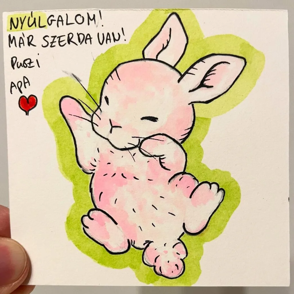 Cute pink bunny outlined in black, lying on its back with one paw raised, surrounded by green highlights and handwritten Hungarian text.
