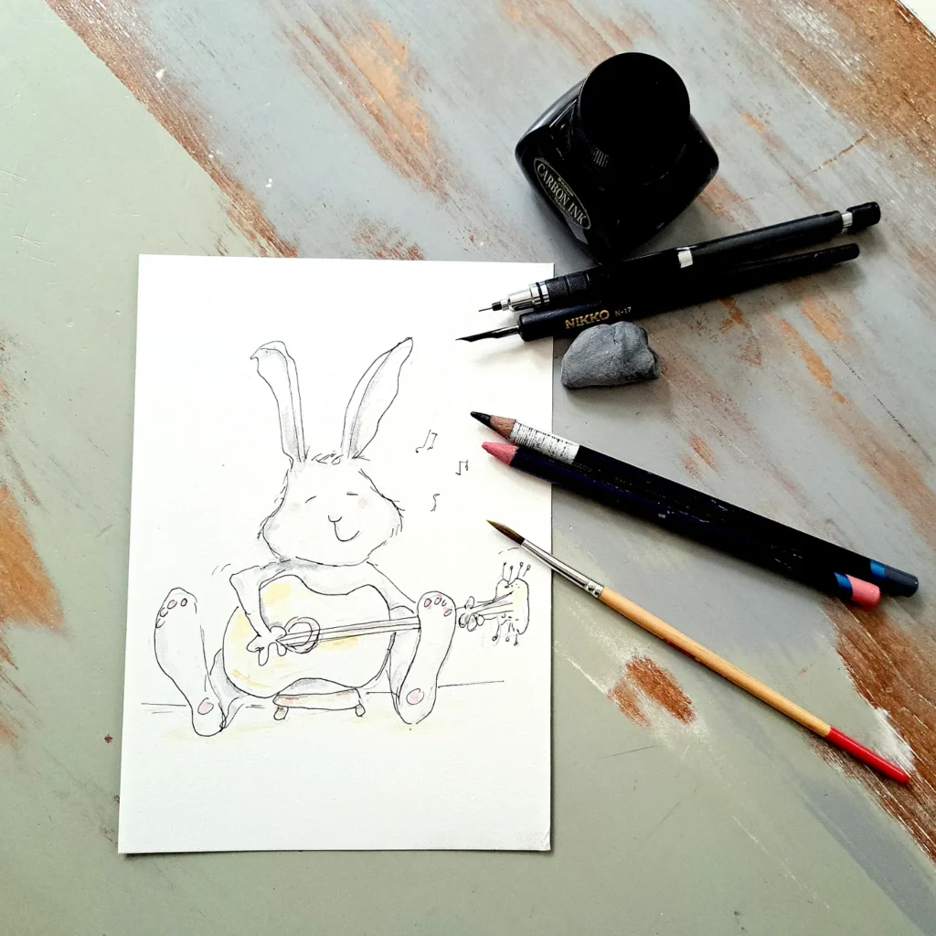 Sketch of a cheerful bunny sitting down and playing a guitar, with art supplies and ink pens visible around the drawing.